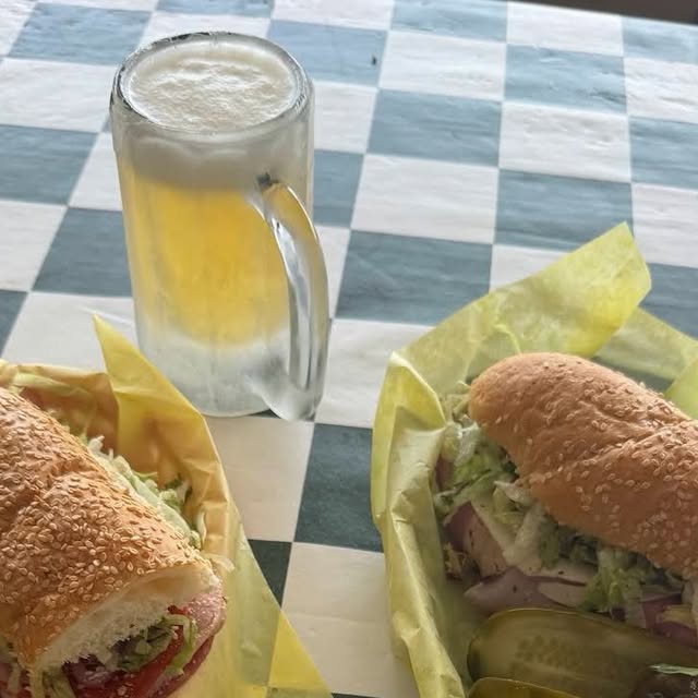Two Dino's subs with pickles and a frosted beer on a green-and-white checkerboard table